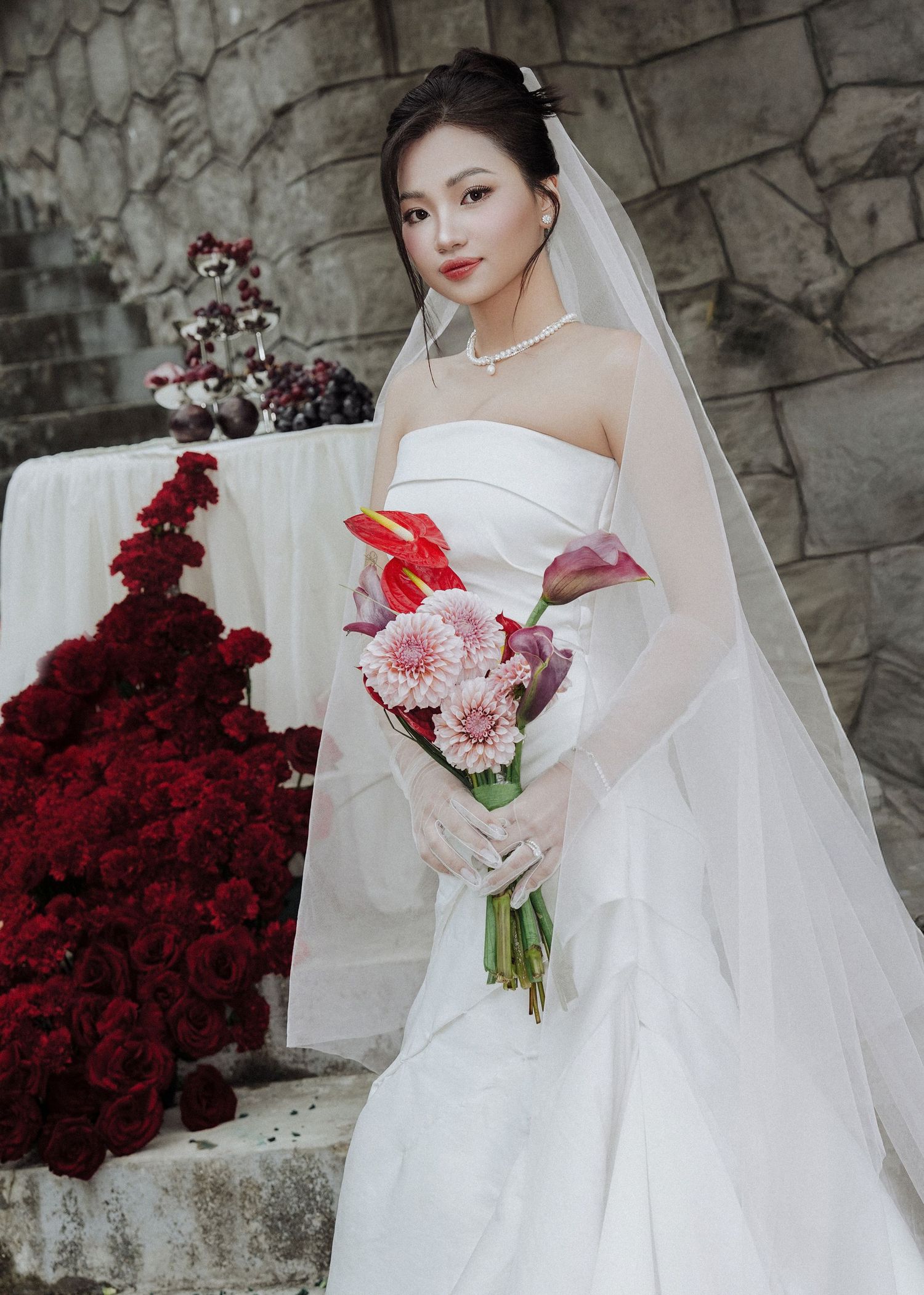 Elegant bride in strapless white gown with veil holding colorful bouquet at stone building backdrop.