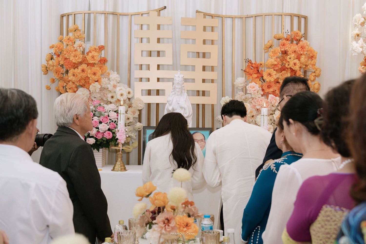 Wedding couple in white ao dai stand before golden cross altar with orange floral arrangements.