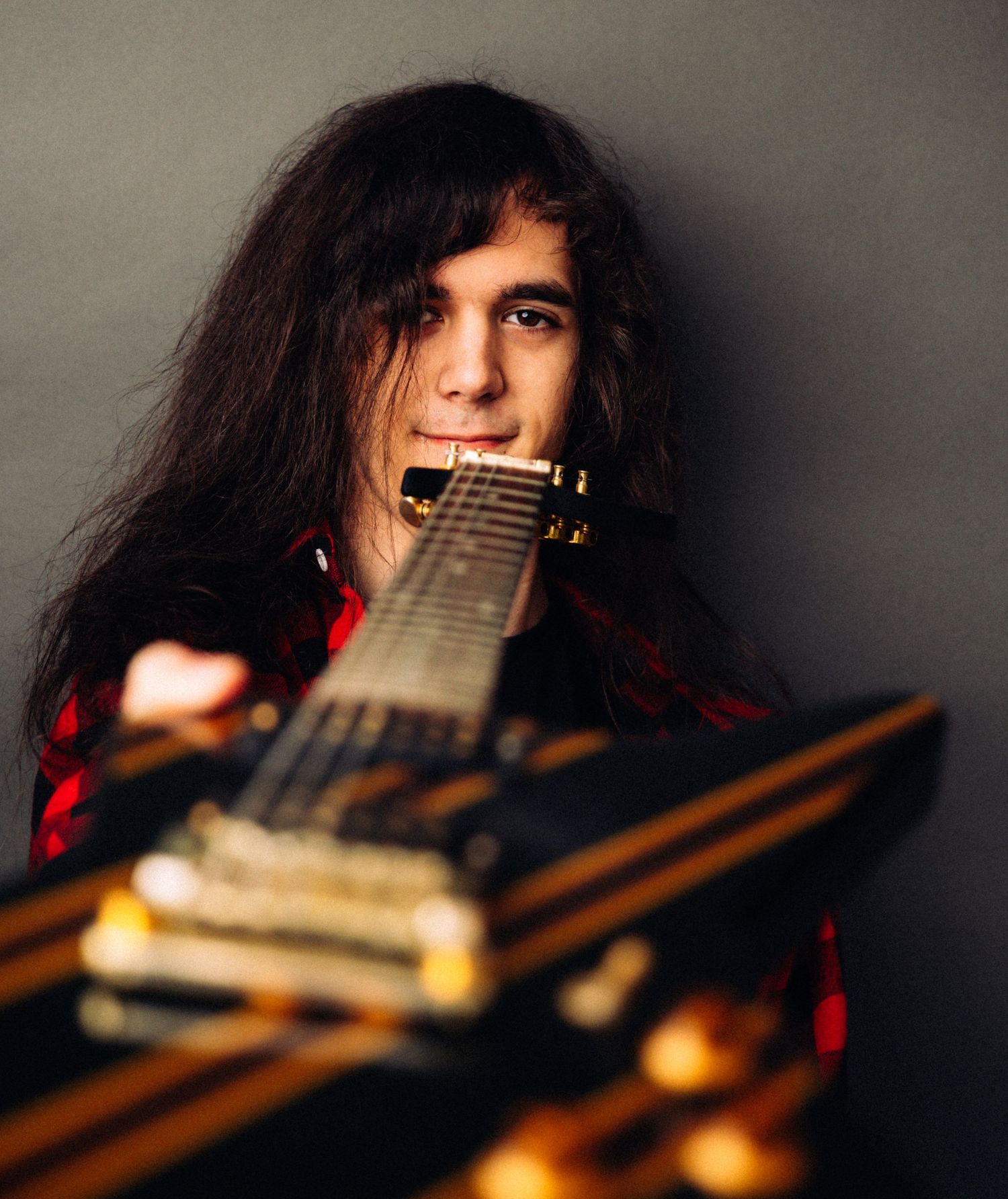 Long-haired guitarist posing with electric guitar against gray studio background.