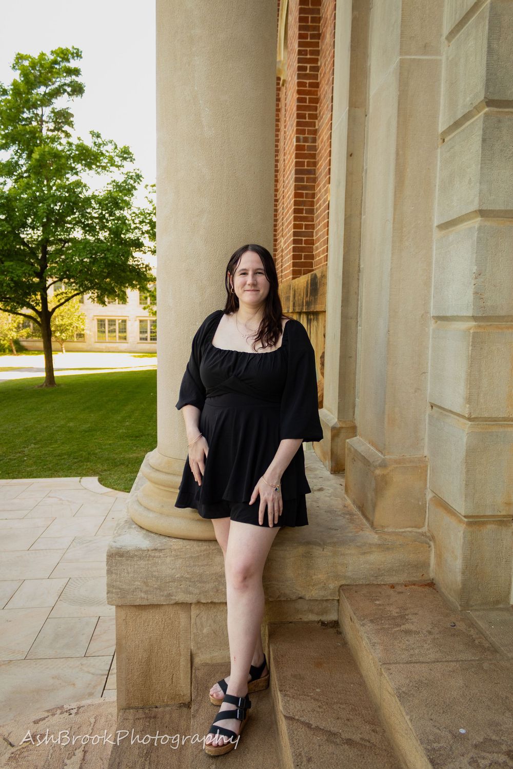 A woman in a black dress, standing in front of a column, hands in a resting position, one foot in front of the other