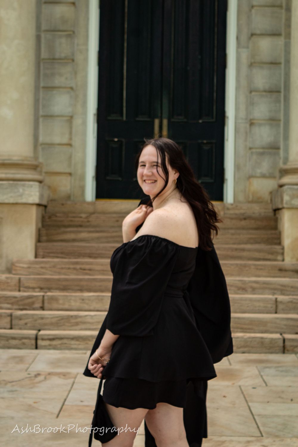 A woman in a black dress looking over her shoulder towards the camera, her graduation gown in one hand, her other hand empty.