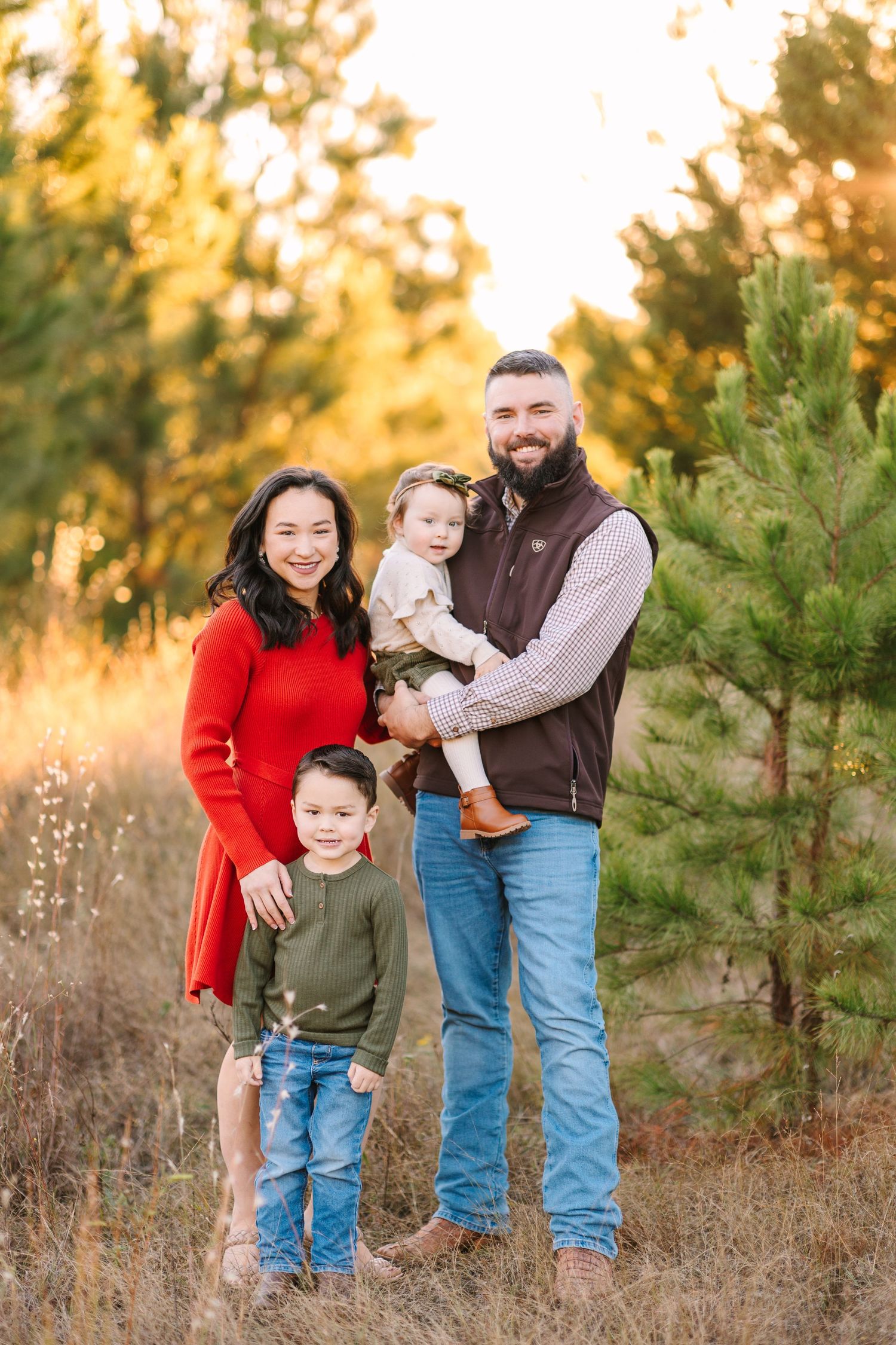 Beautiful fall outdoor portrait shows a family of four standing together among pine trees at golden hour.
