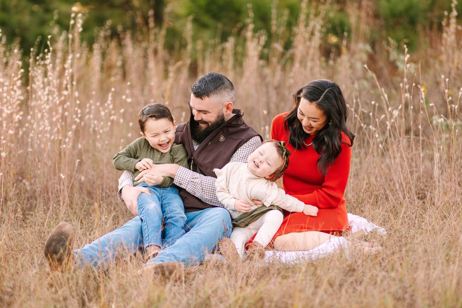 A family lounges together in a sunlit field of tall grass during golden hour, creating a cozy outdoor scene.
