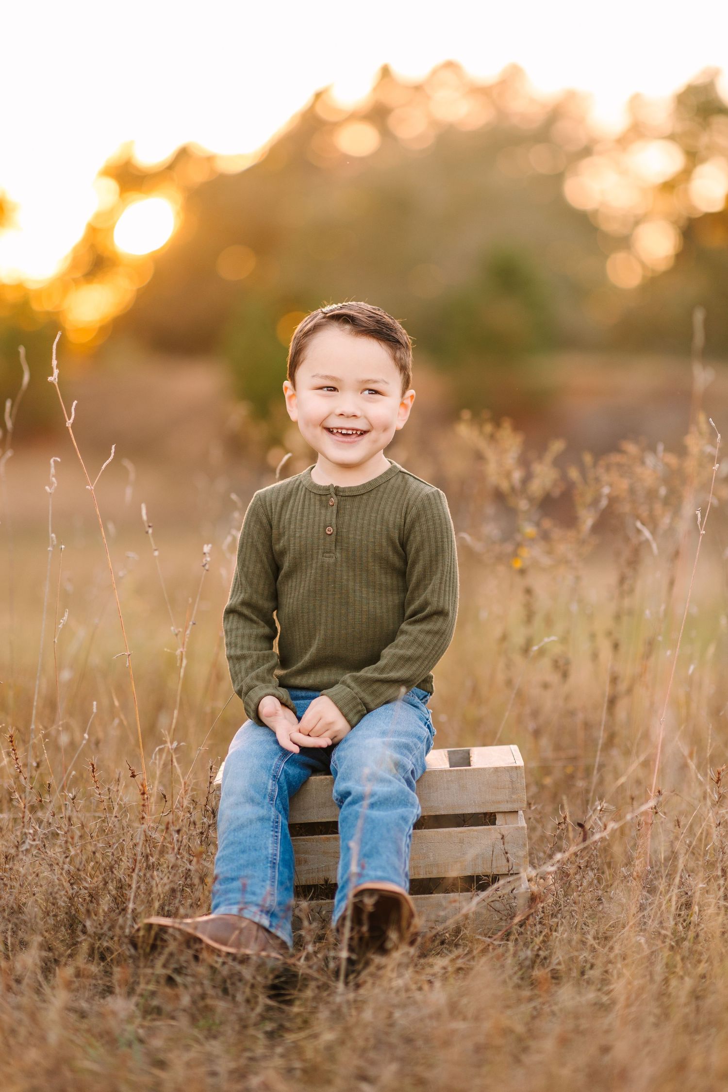 A young child sitting on a wooden crate in a grassy field at golden hour with bokeh lighting behind.