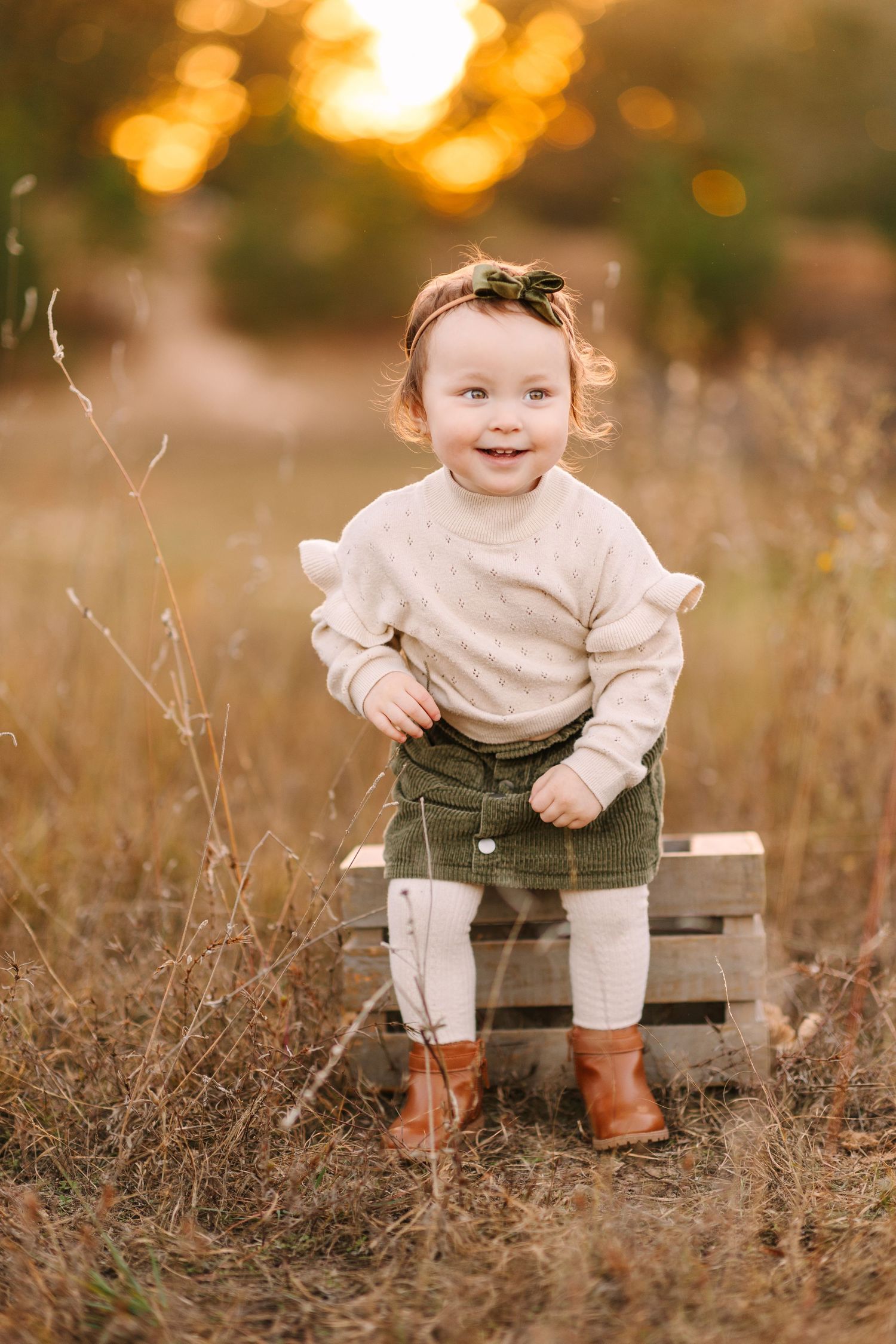 A small child sits on a wooden bench in an outdoor field at golden hour wearing a cream sweater and dark pants.