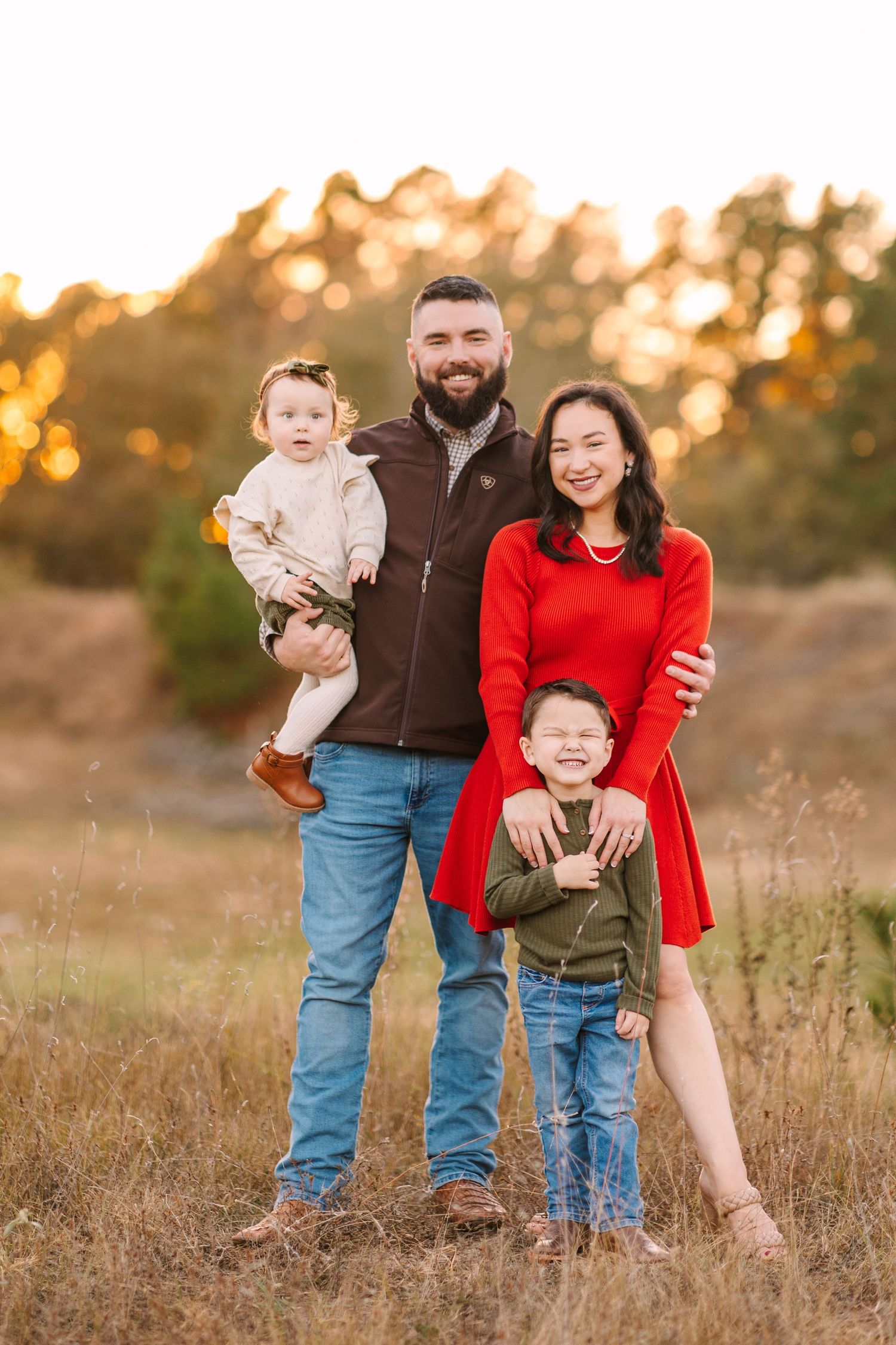 Family poses together in golden field at sunset wearing casual attire with red dress and brown vest.