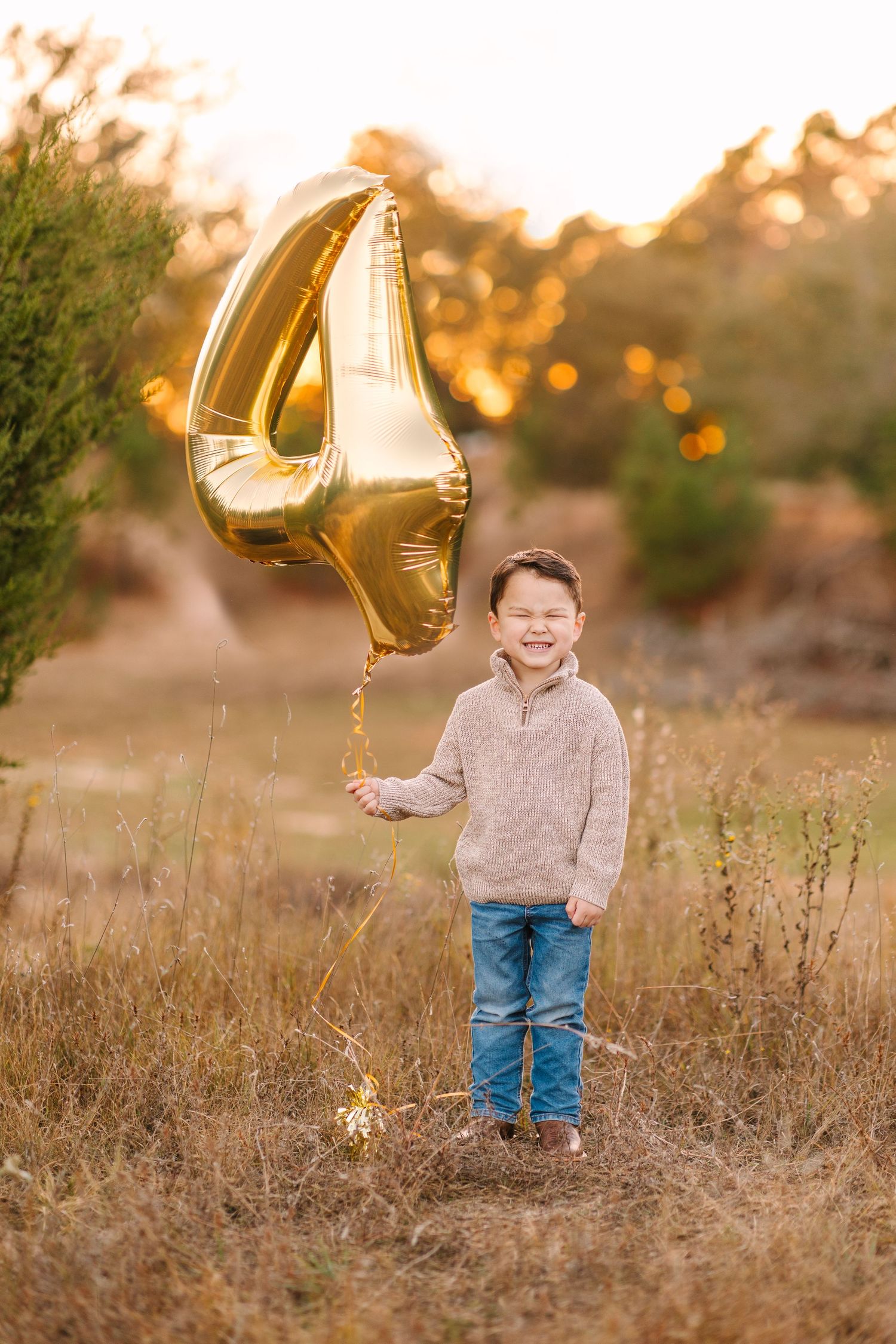 Young child holding a golden number 4 balloon poses in a field during sunset.