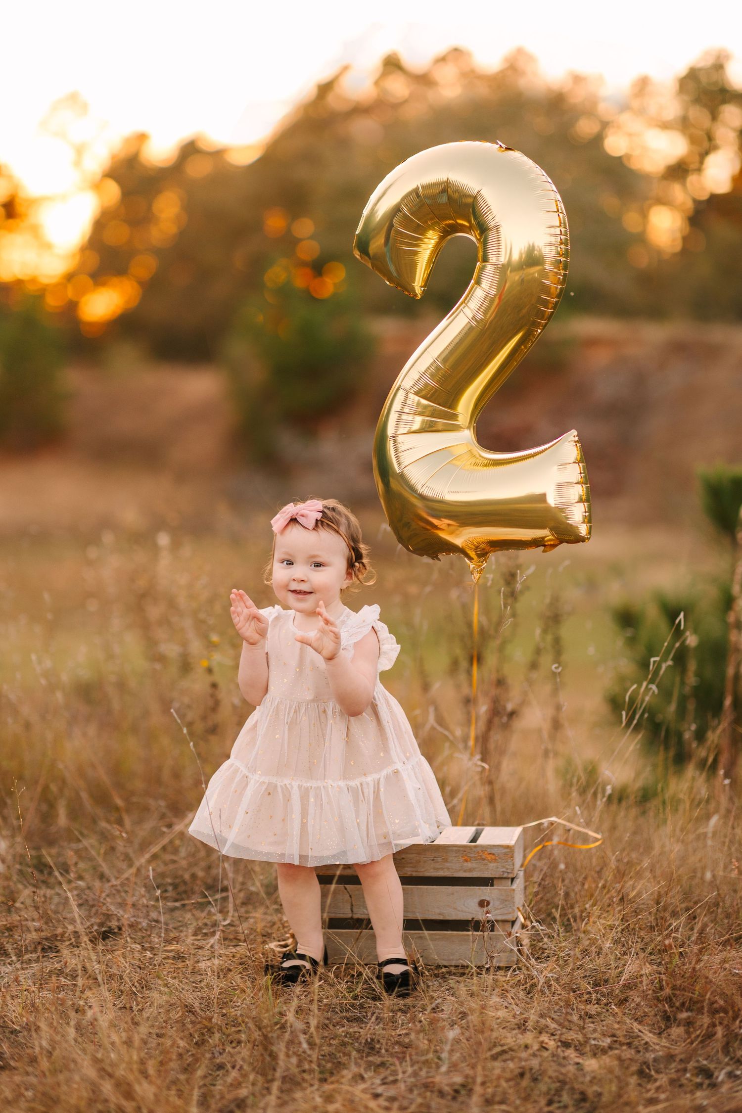 A toddler sits on a wooden crate in a field at sunset with golden number balloons in the background.