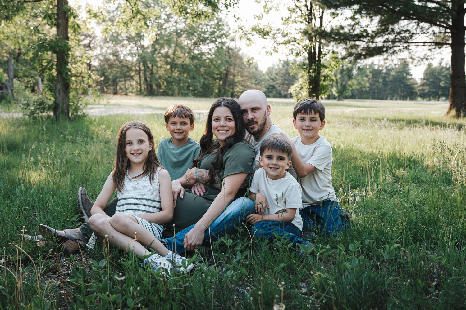 A family group sits together on grass in a sunny meadow surrounded by trees.