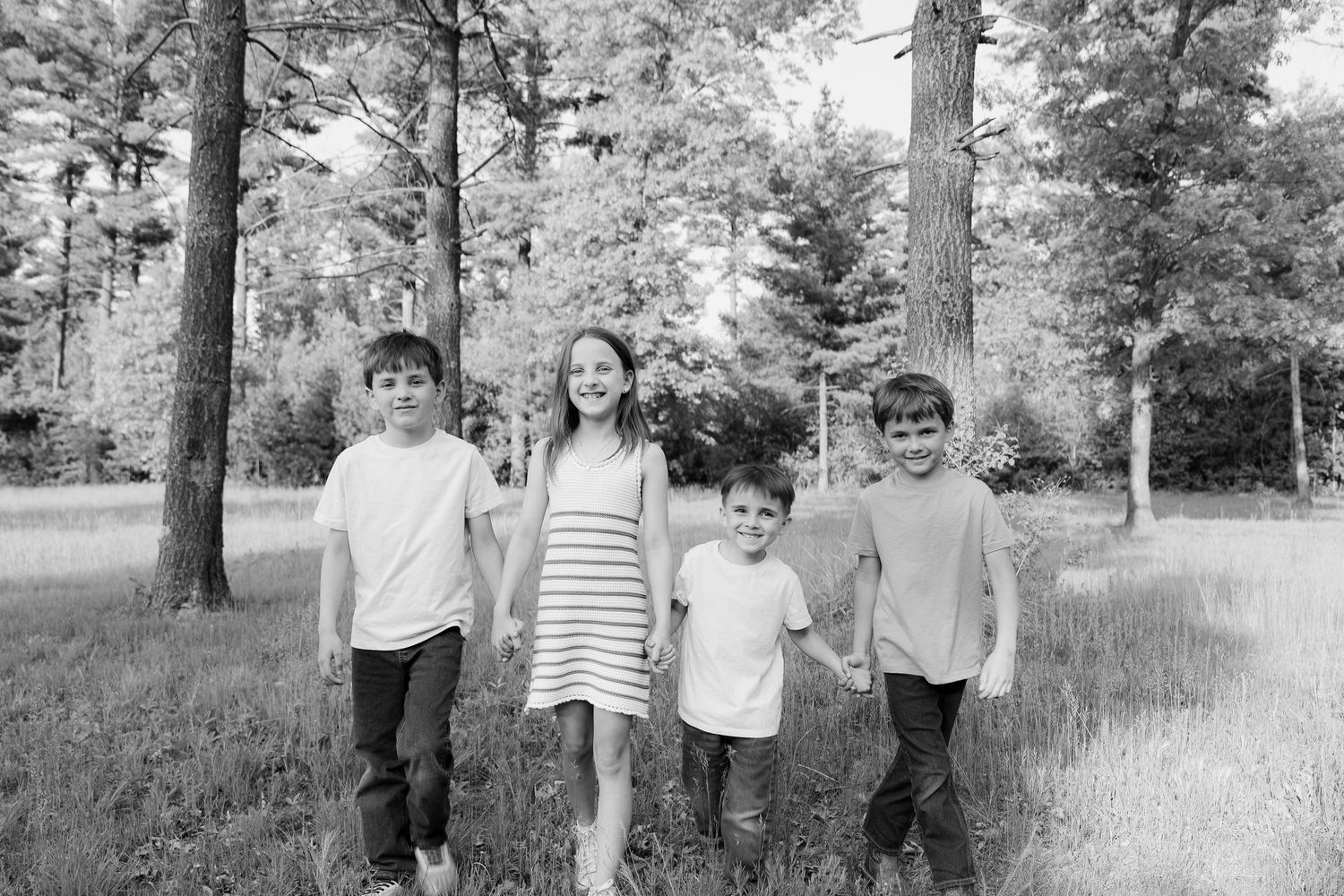 Four children stand together in a pine forest clearing for a black and white portrait.