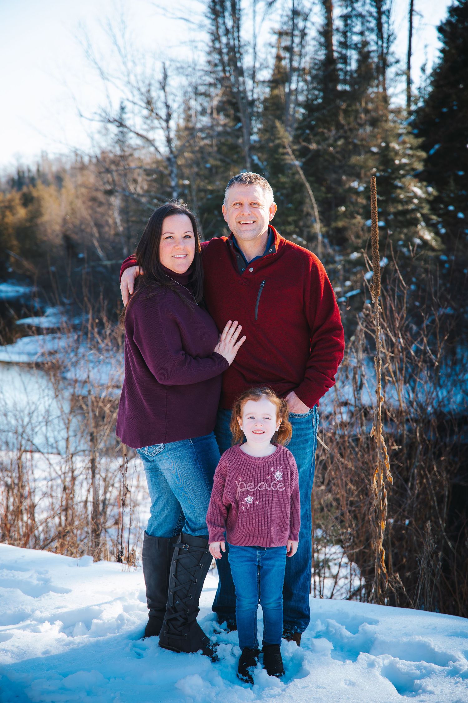 A family of three poses together in winter attire against a snowy forest backdrop.