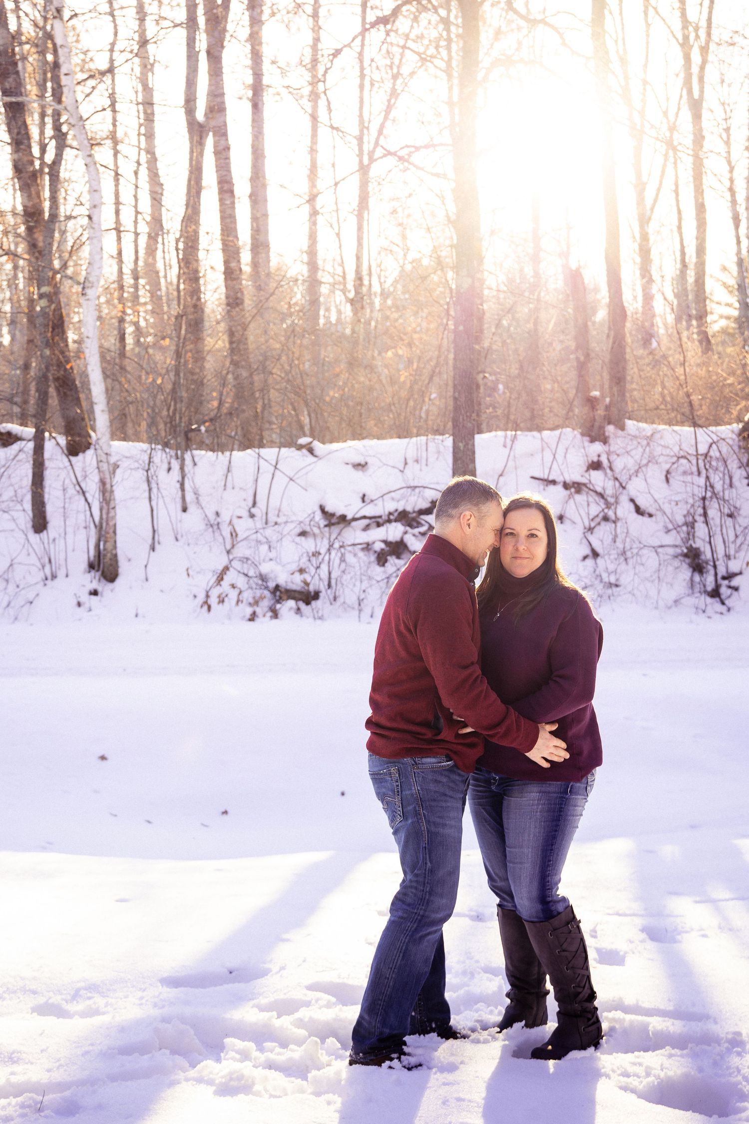 A couple embraces in a sunlit winter forest scene with snow-covered ground.