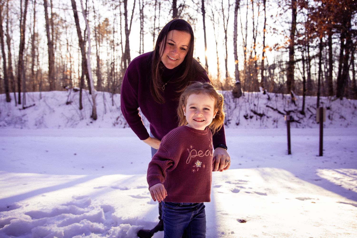 A parent and child share a playful moment in the snow with winter sunlight streaming through trees.