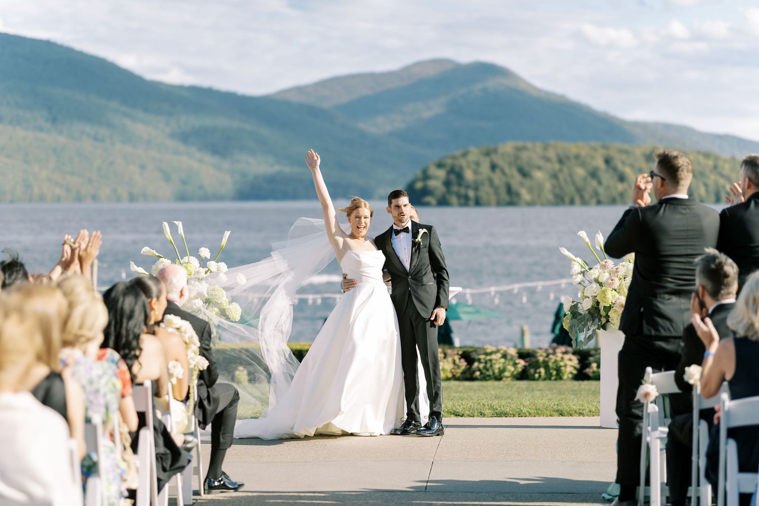 People celebrate during an outdoor wedding ceremony against a backdrop of scenic mountain views and lake.