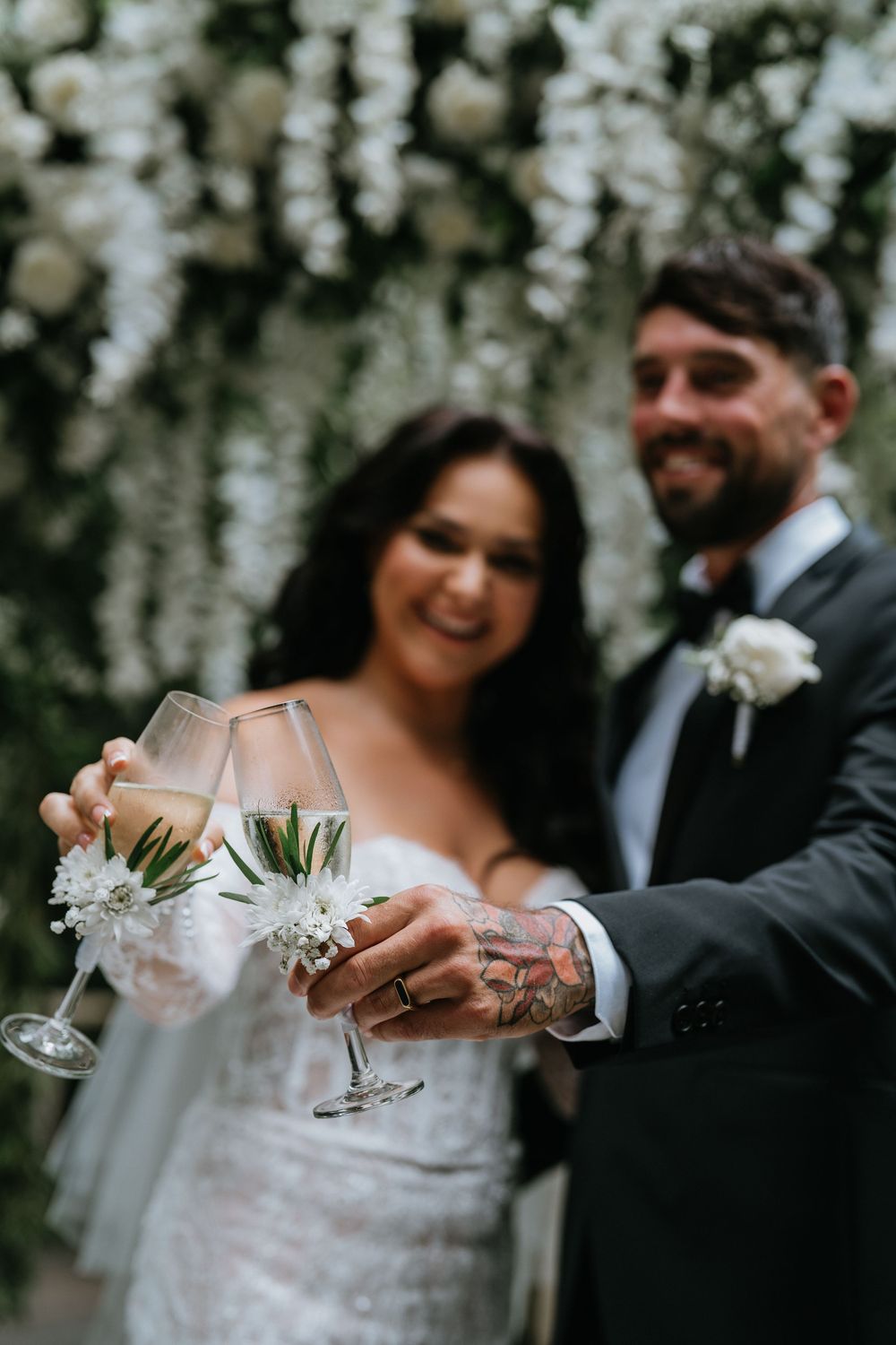 Couple toasts with champagne glasses under white floral wedding decorations.