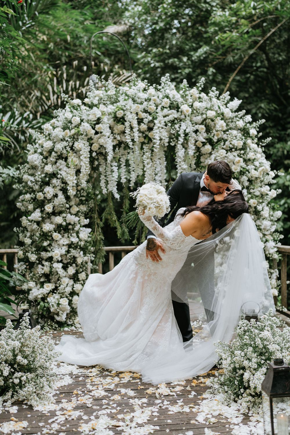 A couple dancing beneath a stunning white floral arch in a garden setting.
