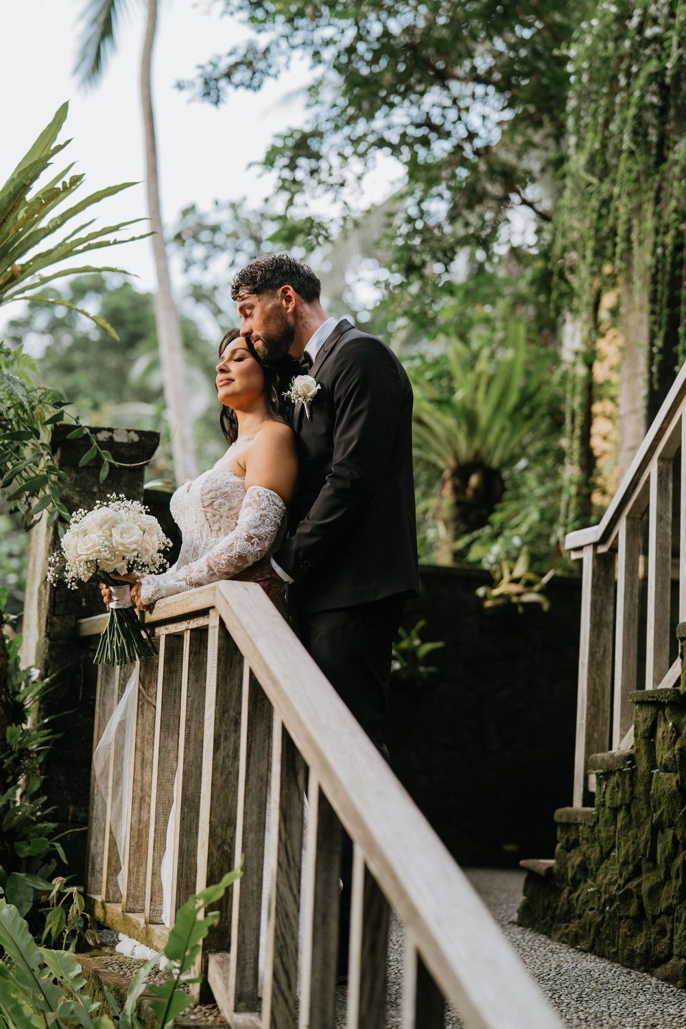 A couple sharing a romantic moment on rustic wooden stairs surrounded by tropical foliage.