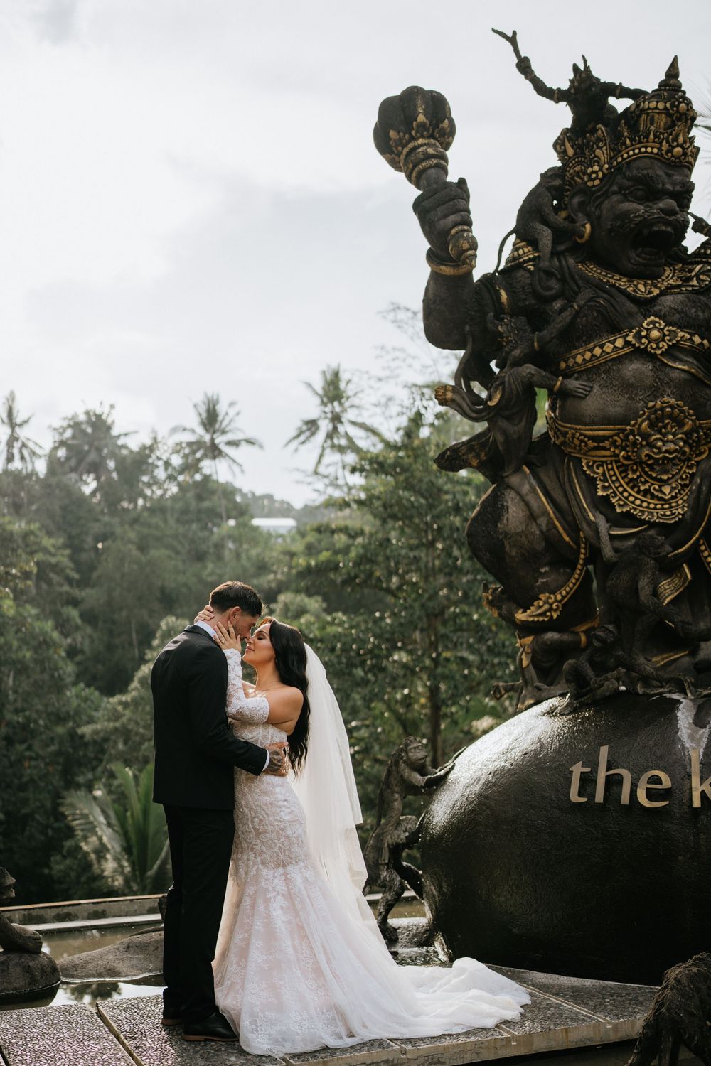 Couple shares romantic moment in front of ornate stone statues and palm trees during outdoor wedding ceremony.