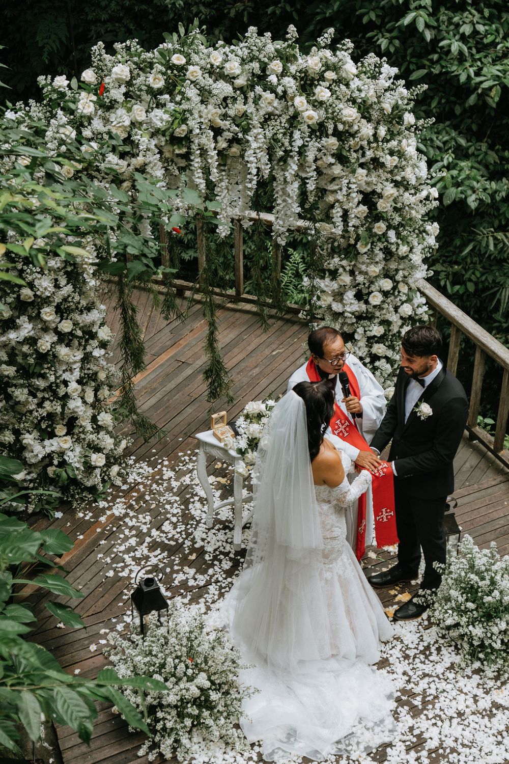 Wedding ceremony on wooden deck surrounded by lush white floral arch and greenery.