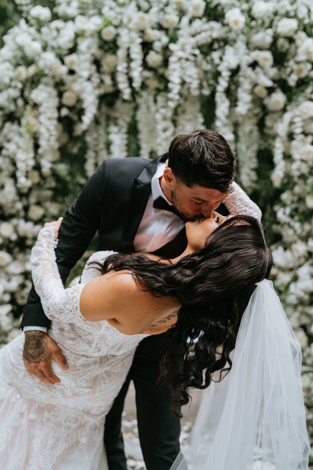 Romantic dip kiss moment under white floral wedding arch during ceremony.