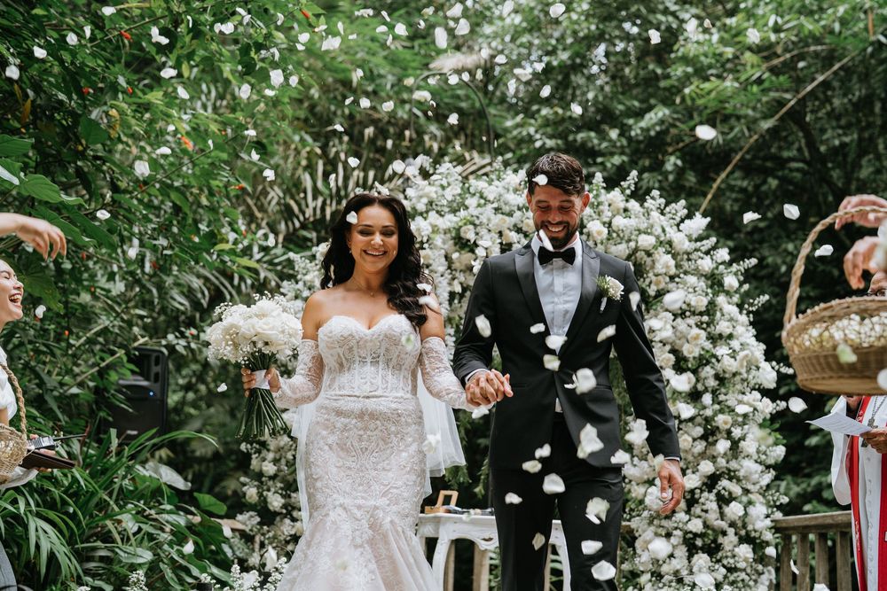 Newlyweds celebrate under cascading white flower arch during outdoor garden ceremony.
