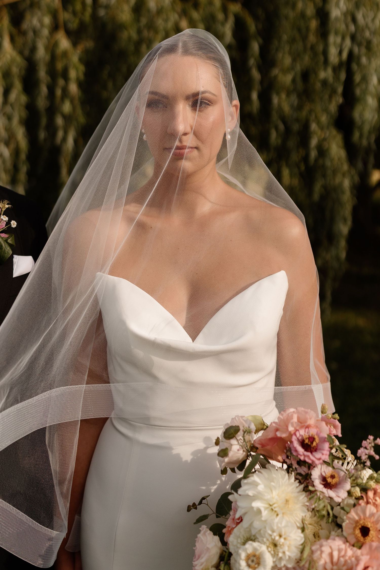 A bride in a strapless white wedding dress holds a soft pink and white bouquet against a backdrop of willow trees. Aliza Dworkind Luxury