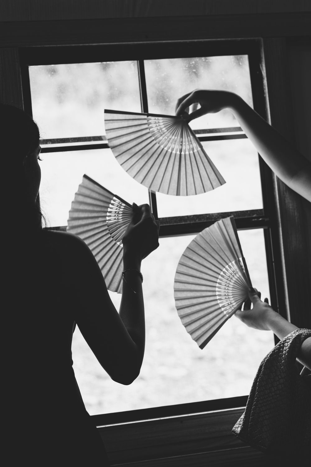 Black and white photo of two people holding decorative hand fans near a window.