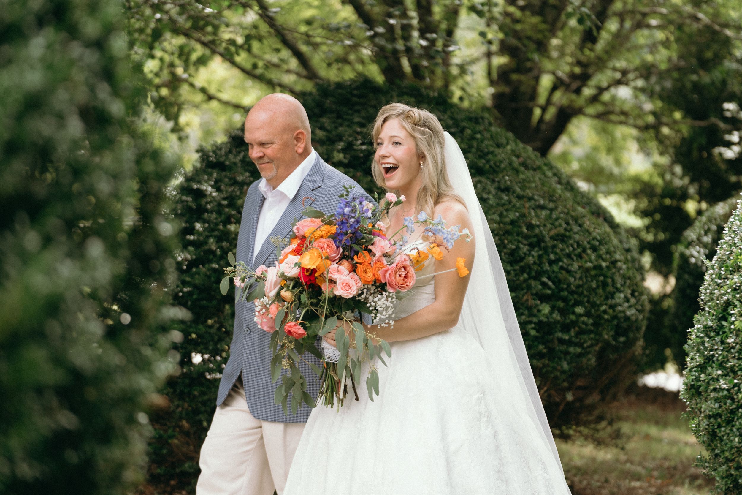 A bride in a white gown holds a vibrant orange and pink bouquet while walking outdoors with someone in a light gray suit. East Coast wedding