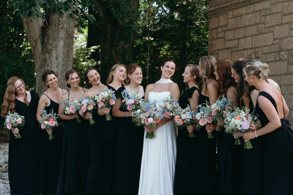 Bride in white gown stands with bridesmaids in black dresses holding pastel floral bouquets outdoors.