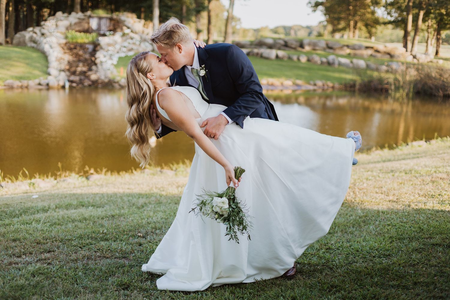 A romantic couple in wedding attire share a tender kiss while sitting by a serene lake on a sunny autumn day.