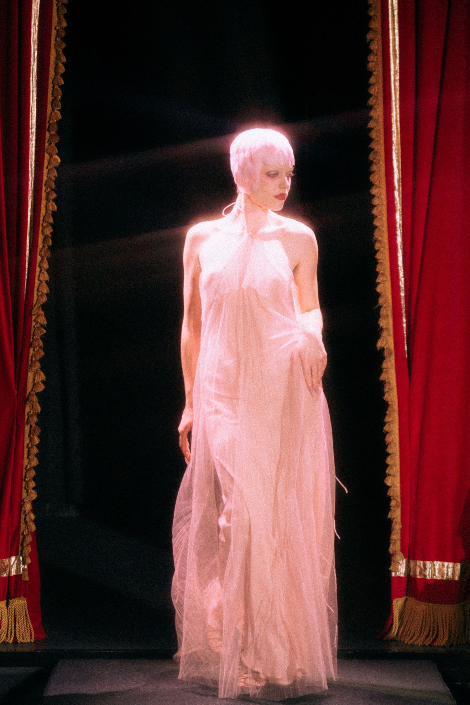 A figure in an ethereal white flowing gown stands dramatically between red velvet theater curtains under stage lighting.