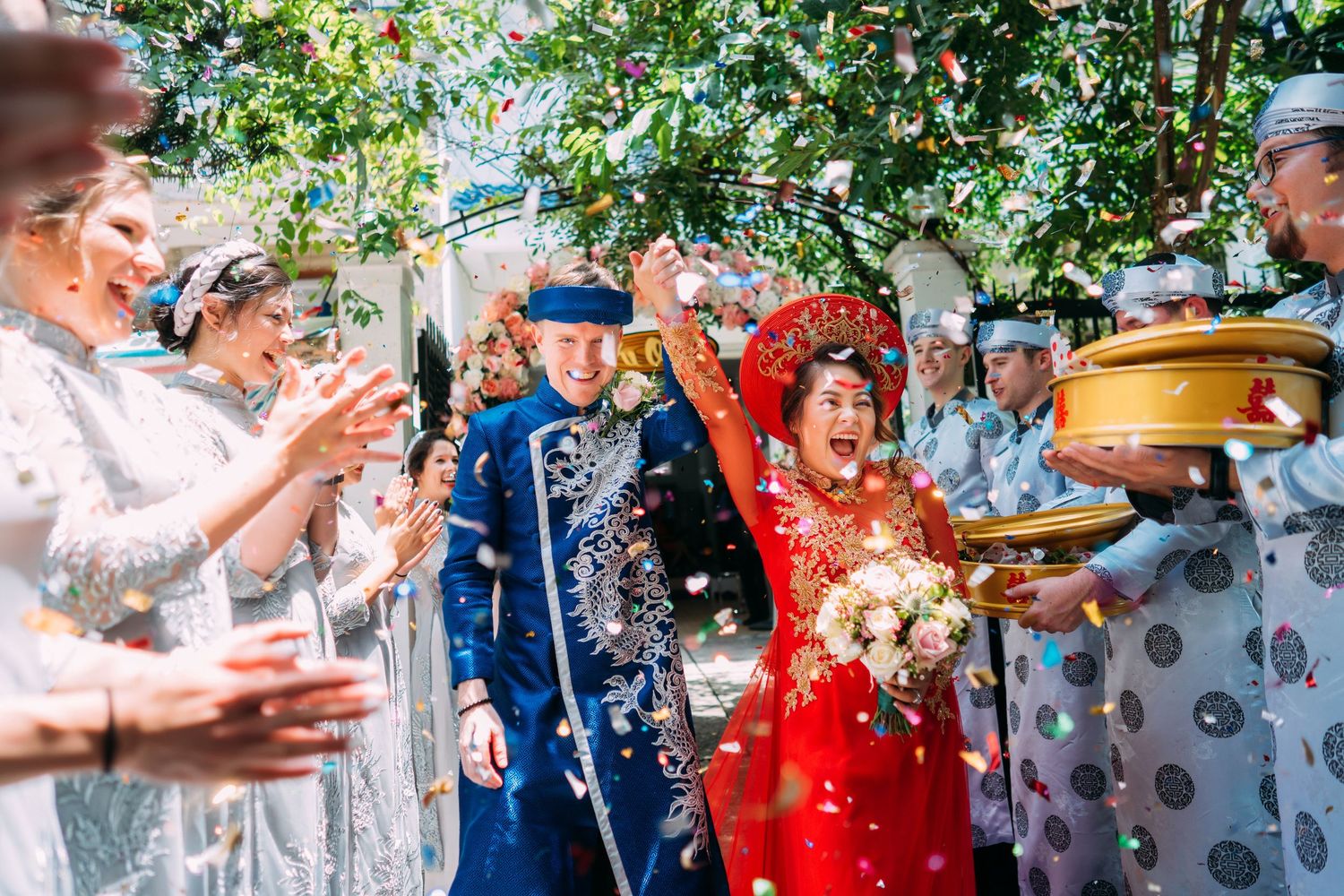 Joyful couple in traditional Vietnamese wedding attire celebrate with confetti shower among cheering guests outdoors.