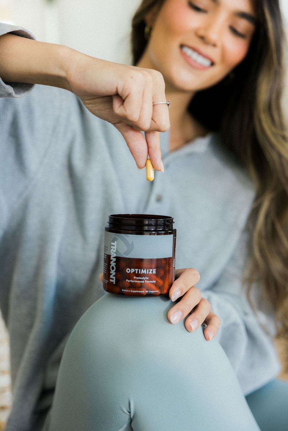 A person in a gray sweater holds and opens a jar of Optimizer supplement product.