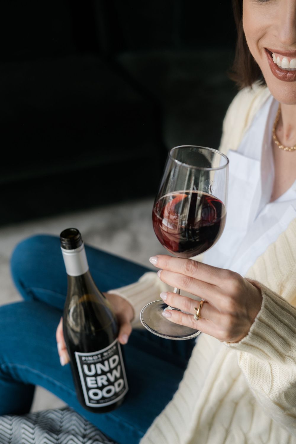 Woman in cream sweater holds wine glass and bottle of red wine while relaxing on blue couch.