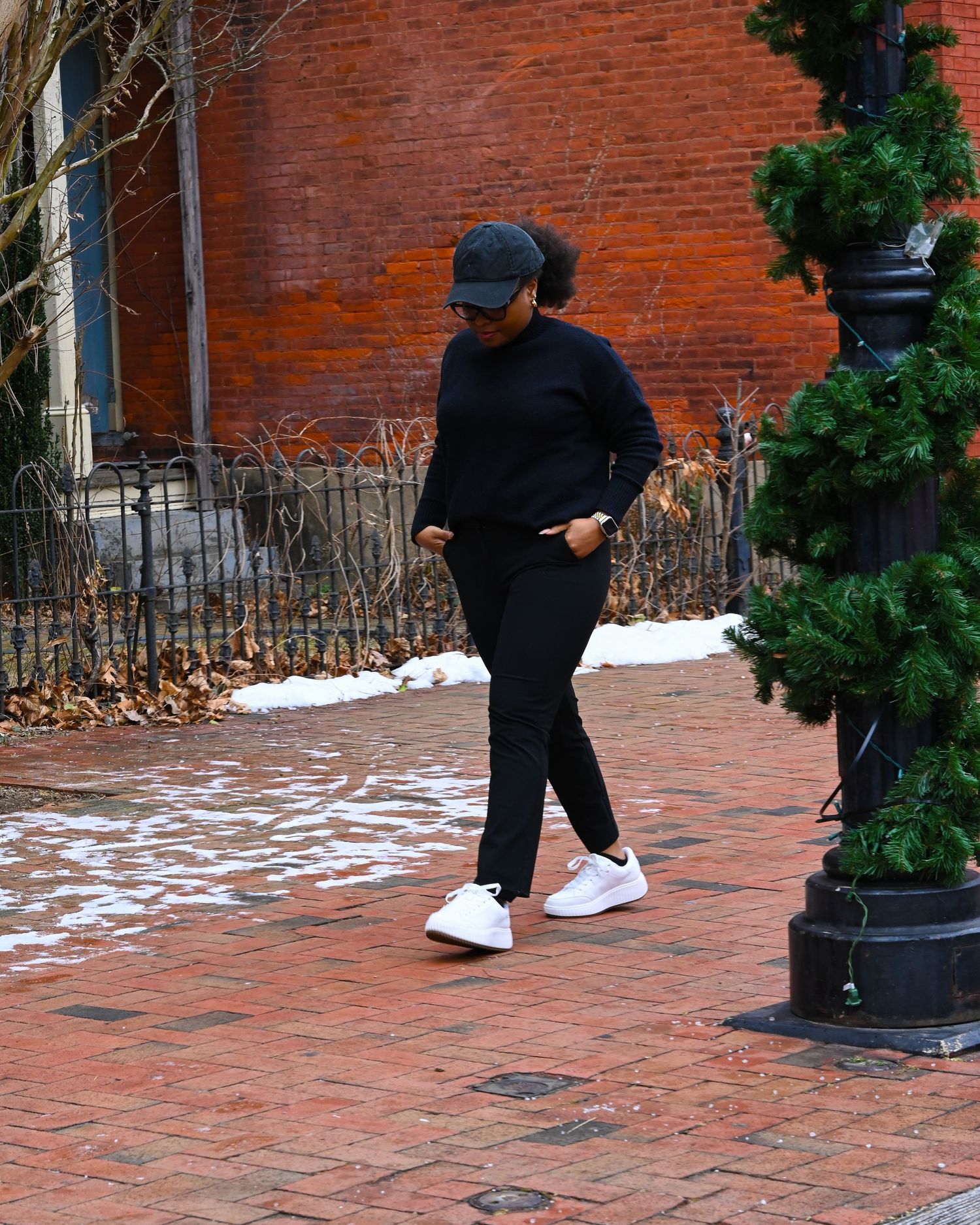 Person in all-black athleisure outfit and baseball cap walking on brick sidewalk during winter with snow patches.