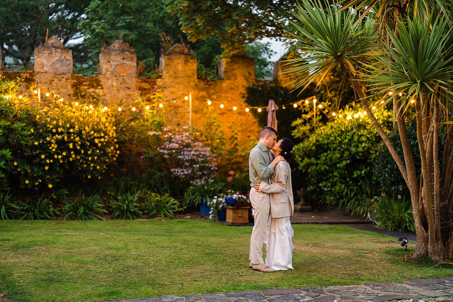 A couple shares an intimate moment in a garden illuminated by string lights at dusk.