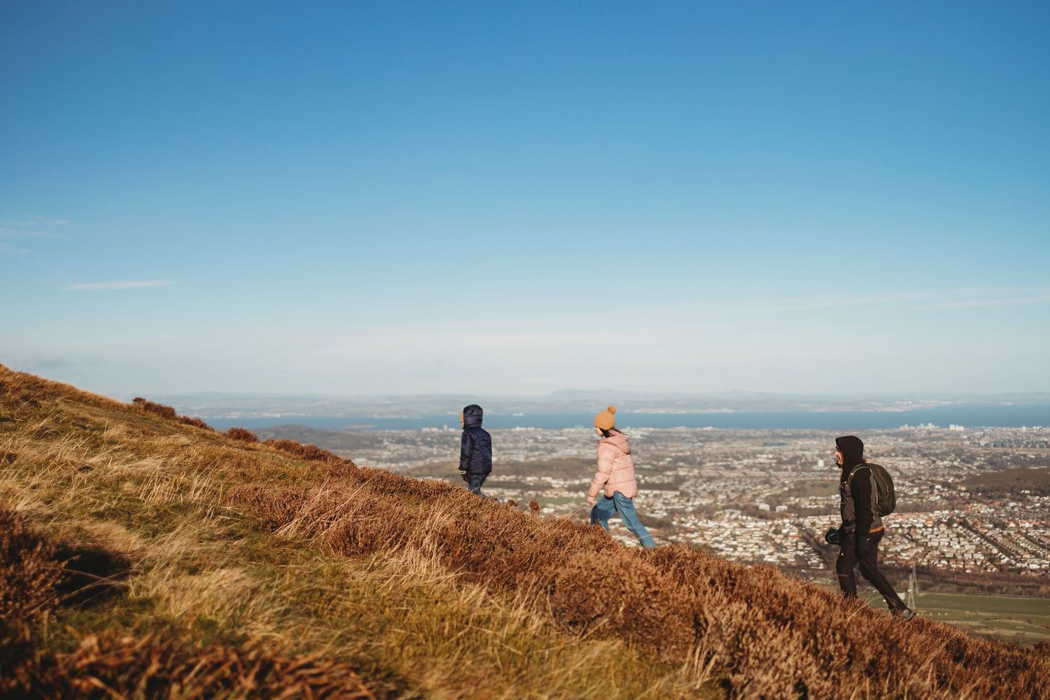 Blue sky stretches over scenic mountain trail with distant city views and hikers on the path.