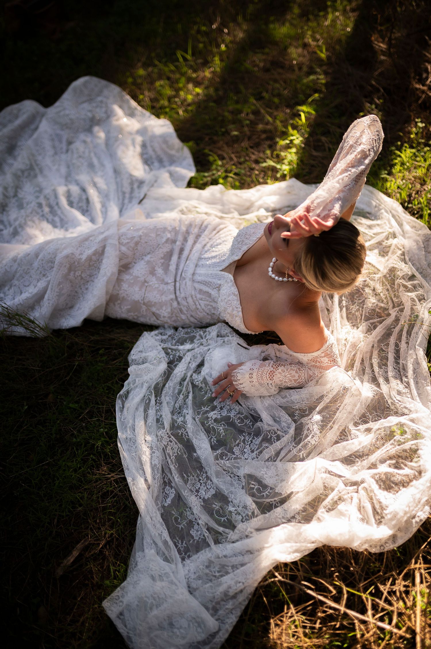 A bride in a lace wedding dress lays gracefully on the ground in dappled sunlight.
