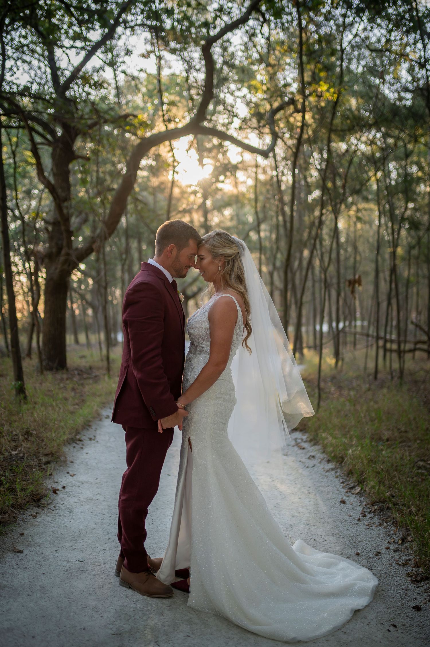 A romantic photoshoot in a wooded area shows a bride in white and groom in burgundy sharing intimate moments at sunset.