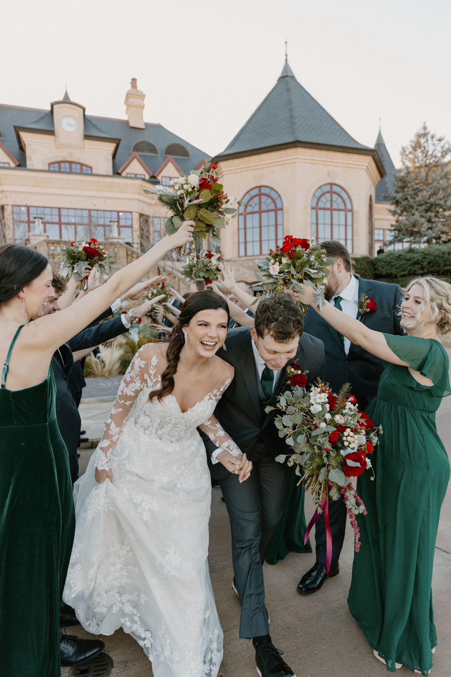 Two newlyweds walk along a curved driveway in front of a grand stone and stucco mansion with arched windows.