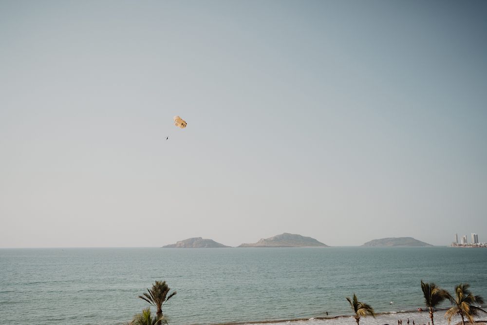 Paraglider soars above tropical beach with palm trees and mountain islands visible on the ocean horizon.