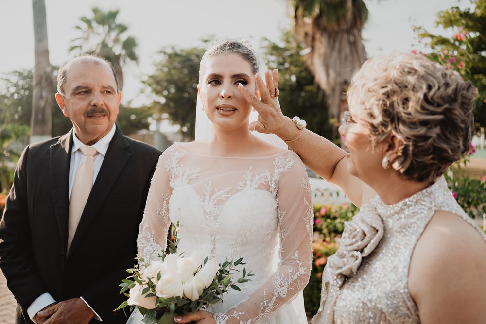A bride in a long-sleeved lace gown stands outdoors with white floral bouquet while being photographed at sunset.