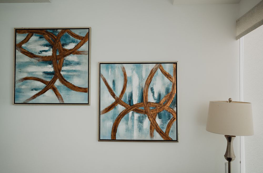 Two modern abstract wall art pieces featuring brown organic lines and curves on turquoise backgrounds hang beside a table lamp.