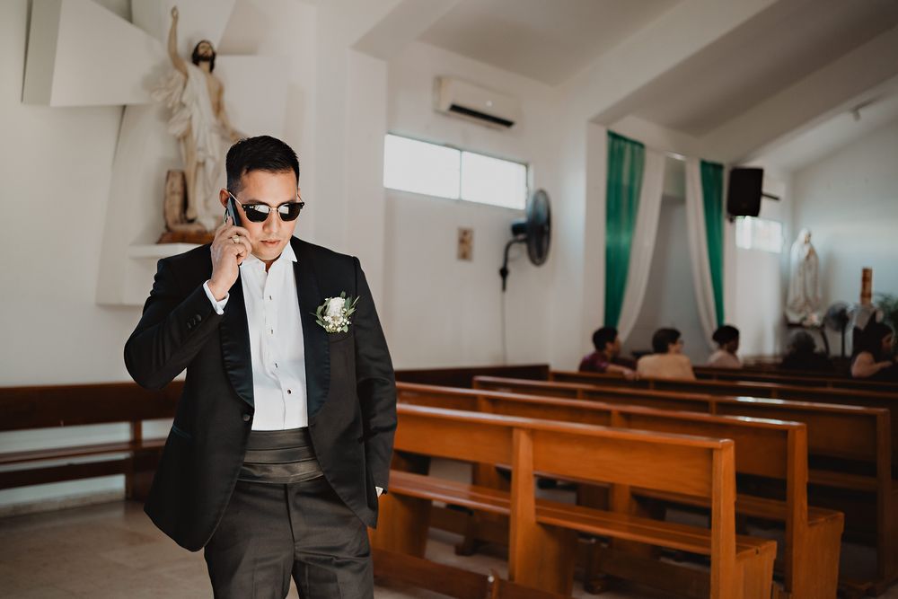 A person in sunglasses and formal black suit stands inside a church with wooden pews and white walls.