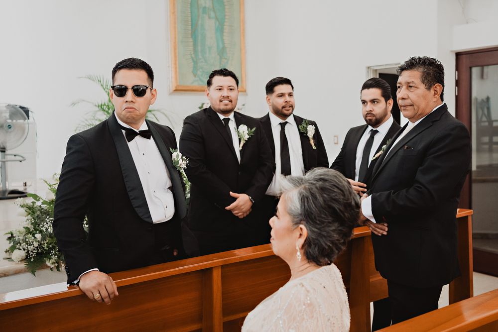 A group in formal attire including black tuxedos and bow ties gather together during a wedding ceremony at a church.