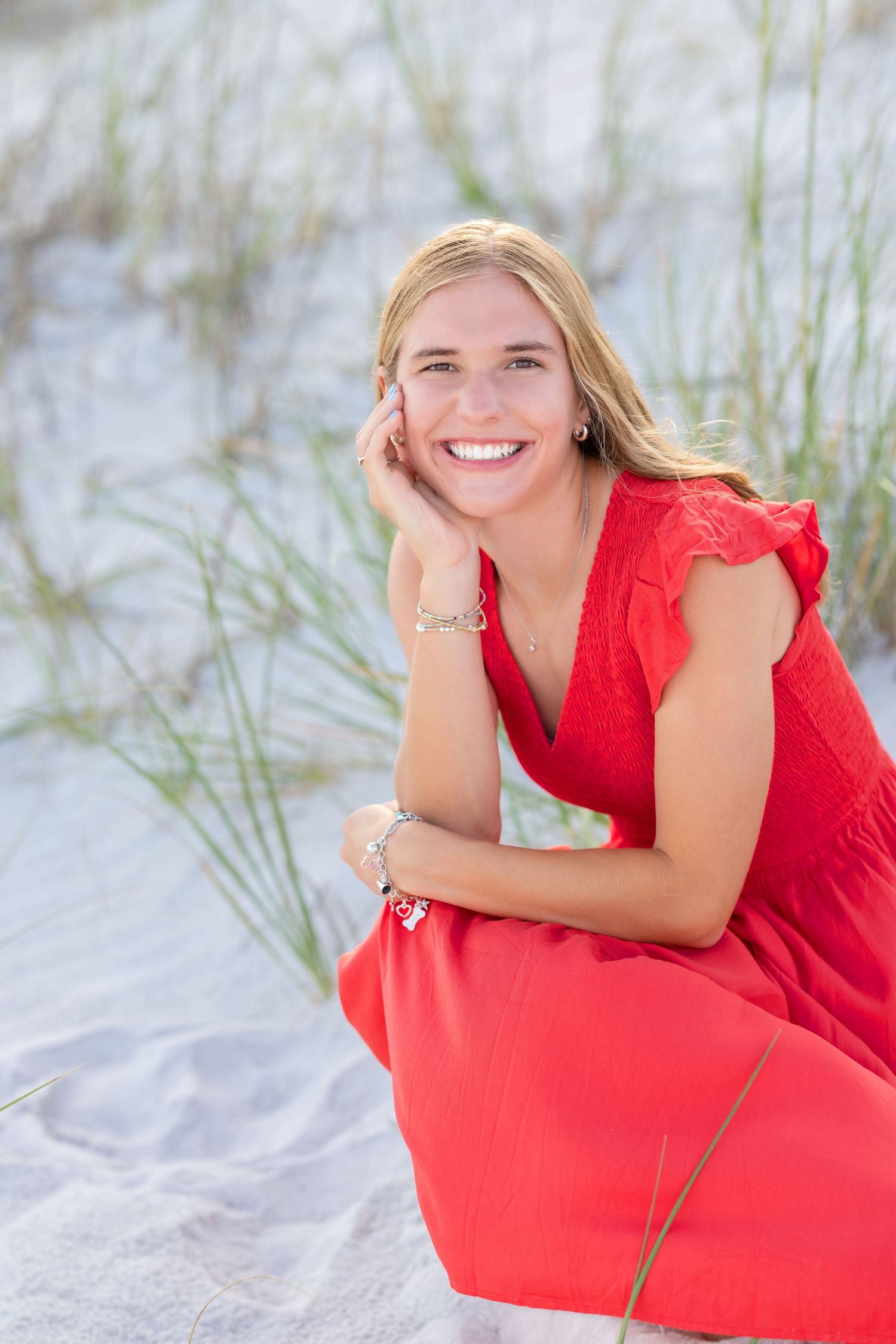 Woman in a vibrant red dress smiles brightly while sitting on a sandy beach with tall grass in the background.