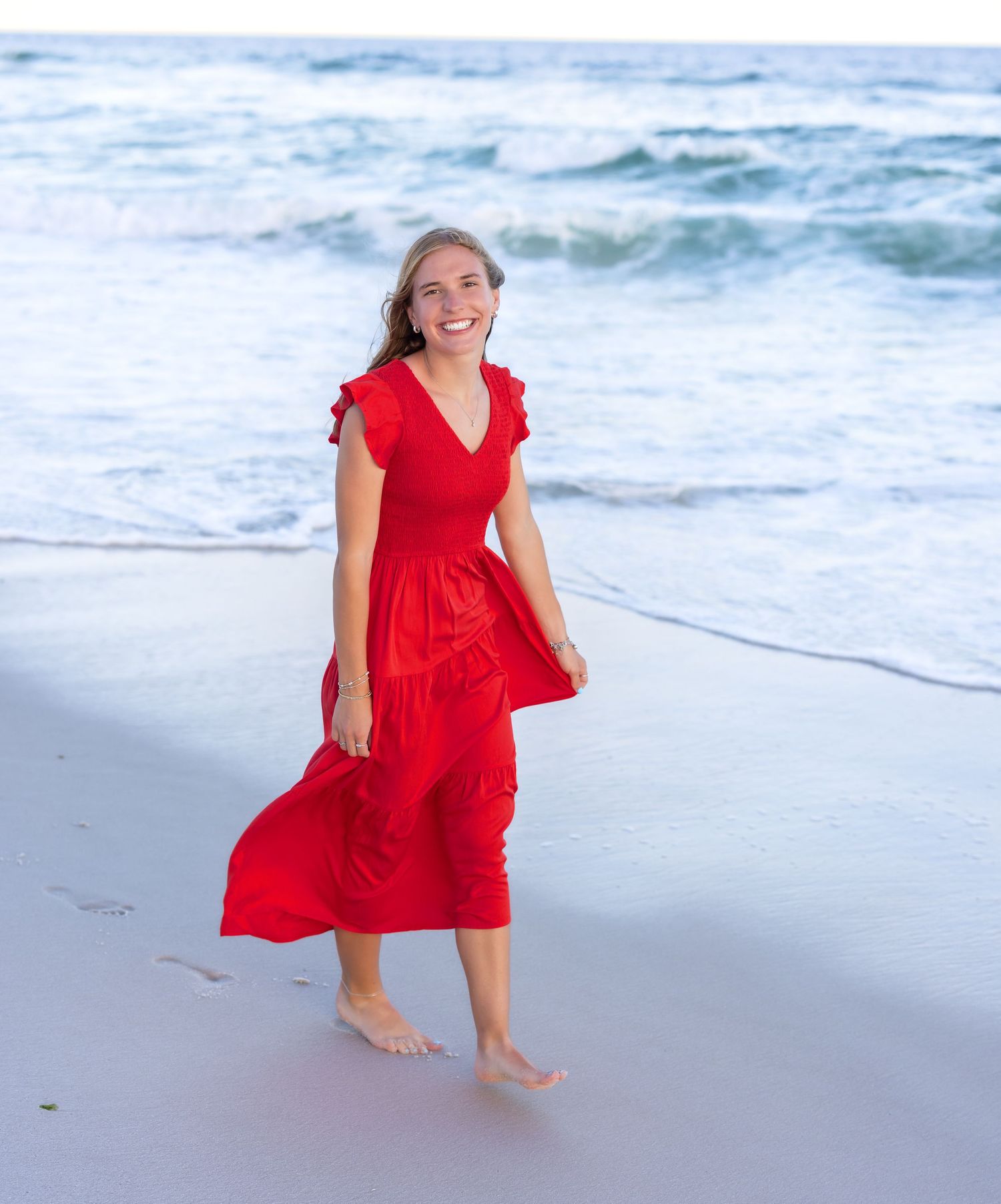 A person wearing a flowing red dress plays on the beach at the water's edge during a bright sunny day.