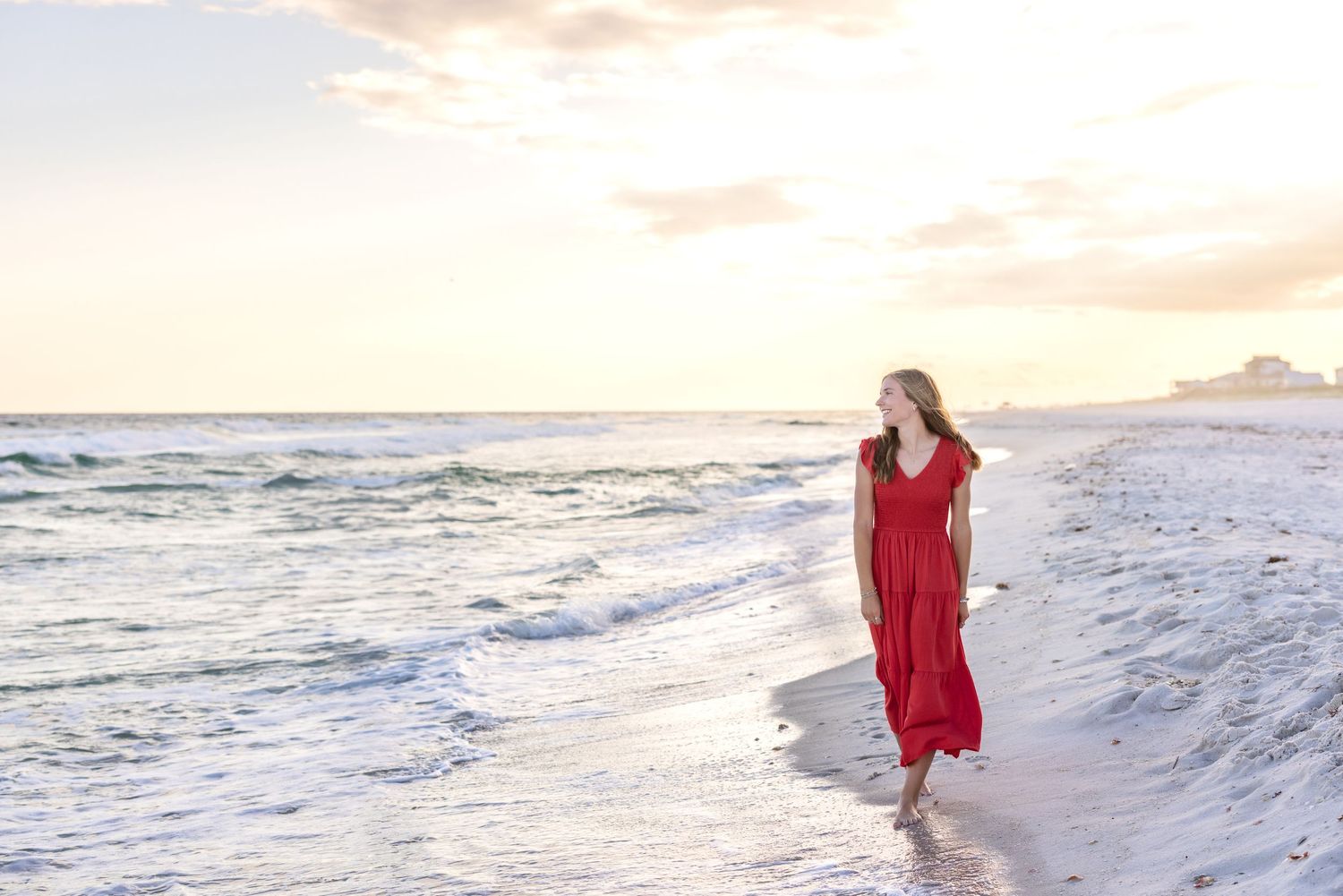 A figure in a flowing red dress walks along a white sandy beach at sunset with gentle waves rolling in.
