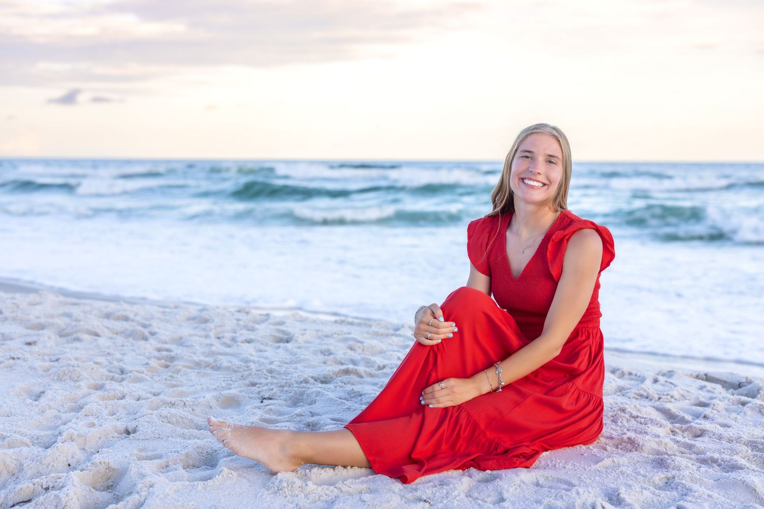 Someone in a flowing red dress poses gracefully on a sandy beach at sunset with ocean waves in the background.