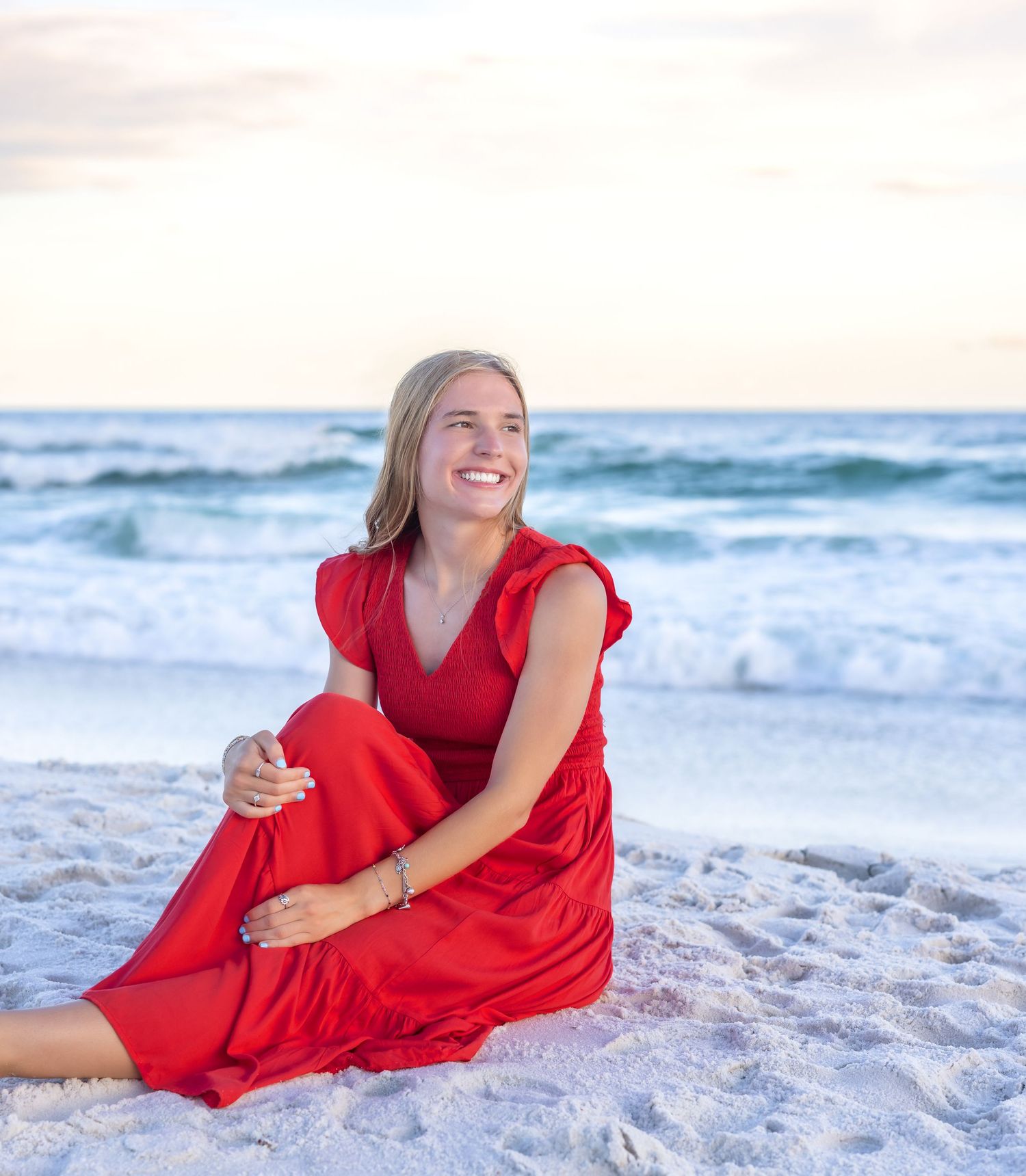 Someone in a flowing red dress sits on a white sandy beach at sunset with ocean waves in the background.
